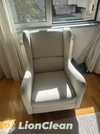 Svetlosiva fotelja sa visokim naslonom i bocnim krilima pored prozora, pogled spreda